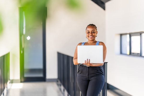 African black business woman holding a laptop at office. Smiling young African businesswoman looking up while working on laptop.