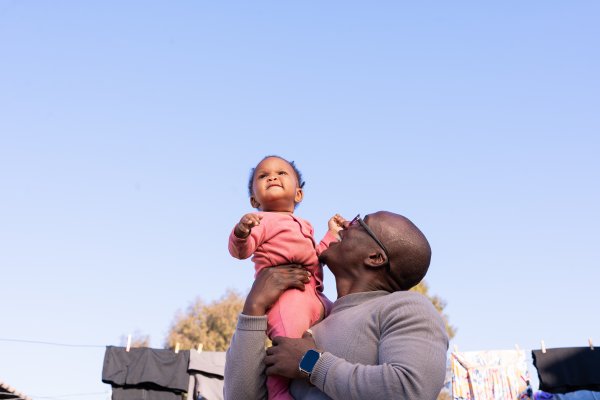 Black father smiling outdoors with daughter wearing pink and blue sky.