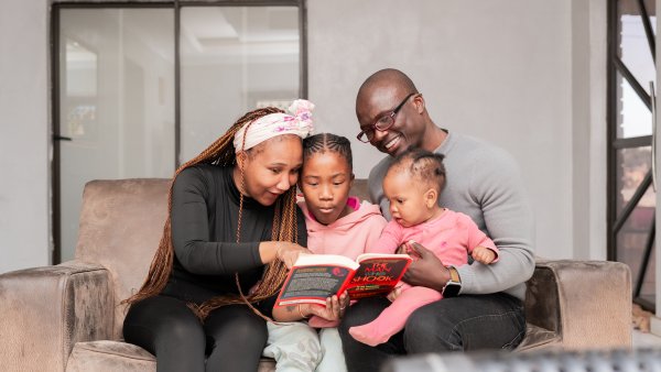 Loving black family reading a book at home on the couch, mom wearing black and kids wearing pink.