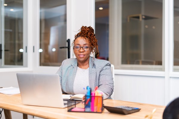 Mature African black woman sitting at desk with laptop and wearing glasses in a co working space.