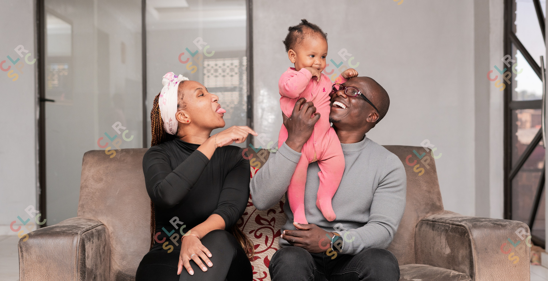 A joyful family smiles at home. Dad, Mom in black, and their 1-year-old daughter in pink.