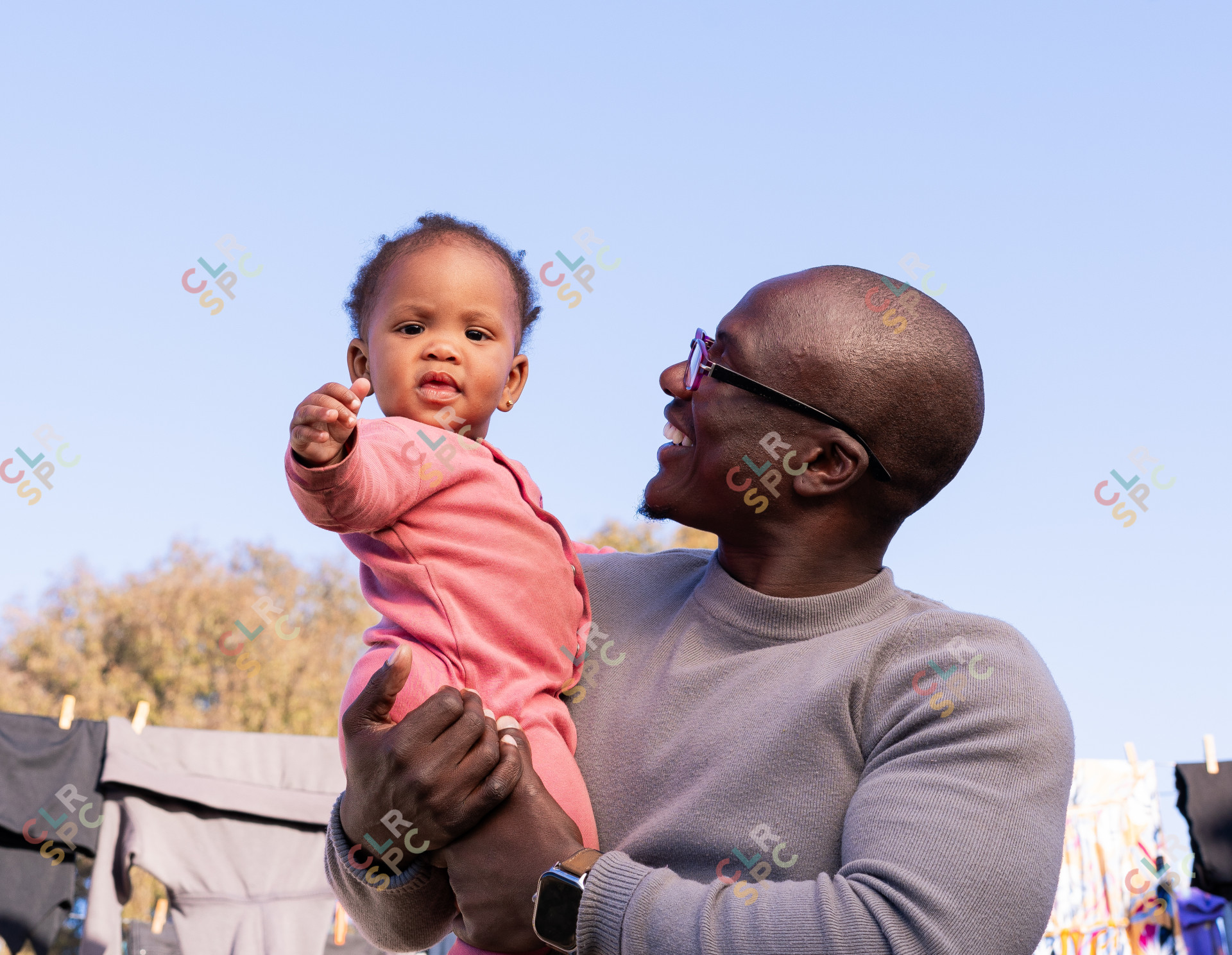 Black father holding daughter and smiling in the sun with clothe hanging in the background.