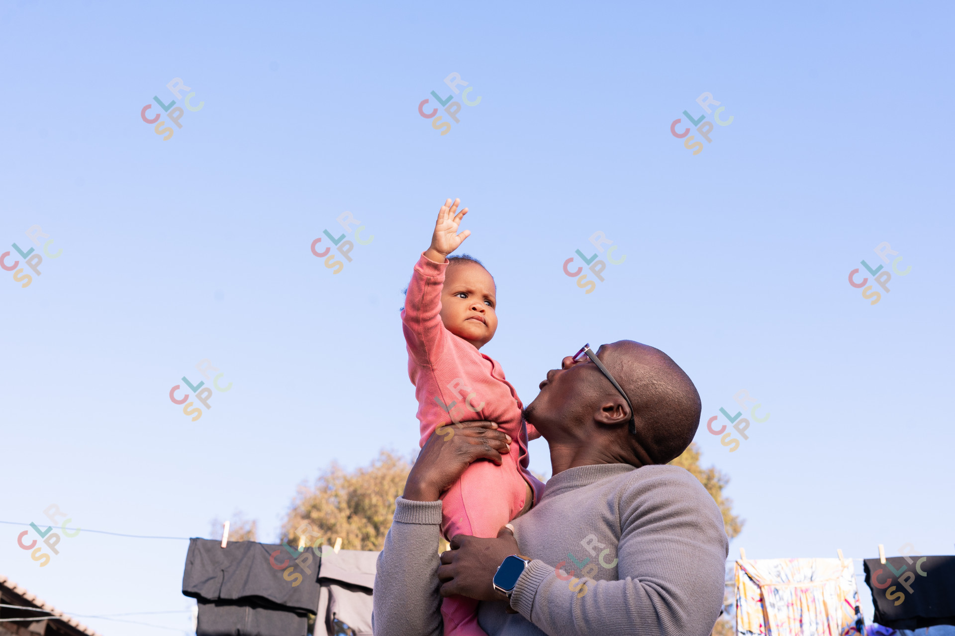 Black father playing outside with daughter wearing pink and clothes in the background with blue sky.