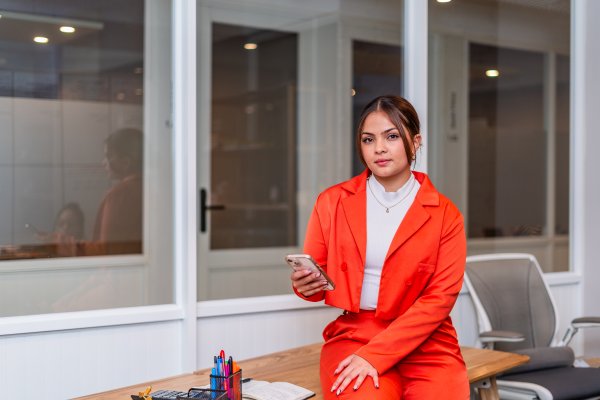 Professional South African woman wearing orange suit at the office holding a phone sitting on a desk.