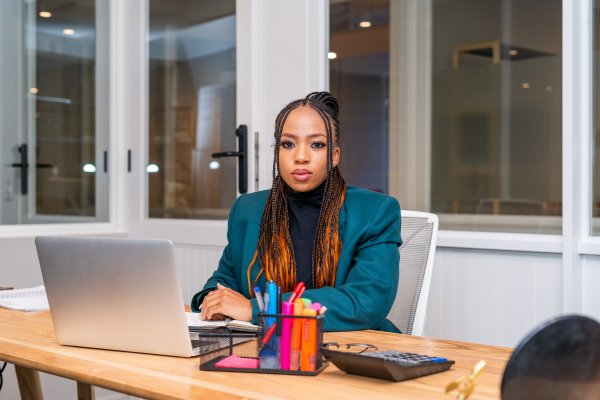 Young black woman sitting at her desk at work looking at the camera and a laptop in front of her.